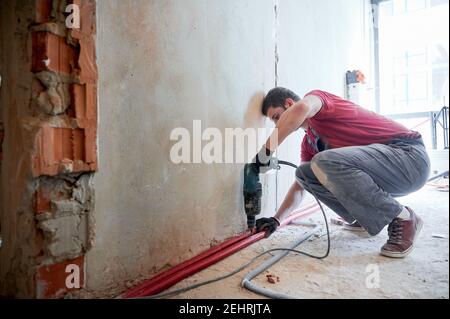 Vue latérale d'un plombier barbu installant un système de chauffage au sol dans une pièce vide avec des murs en briques. Homme portant une combinaison et des gants, debout sur un genou, travaillant avec une perceuse électrique Banque D'Images