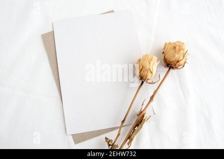 Mode mariage papeterie maquette scène. Une carte de vœux verticale vide et des roses blanches séchées sur un fond de lin blanc. Thème féminin, vue du dessus. Banque D'Images