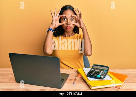 Jeune fille afro-américaine travaillant au bureau avec un ordinateur portable et une calculatrice essayant d'ouvrir les yeux avec les doigts, endormie et fatiguée pour la fatigue matinale Banque D'Images