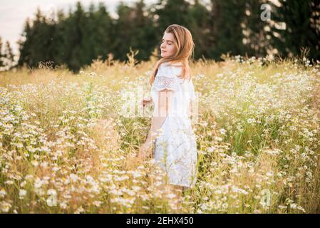 Jeune fille mignonne, tendre rêve amoureux, dans un champ avec des pâquerettes. En robe et chapeau en osier. Chaud été ensoleillé, coucher de soleil dans le village. Concept de liberté et Banque D'Images