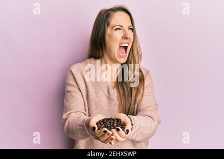 Jeune femme blonde tenant des grains de café avec les mains en colère et fou hurlant frustré et furieux, criant avec colère. Rage et agressif concept. Banque D'Images