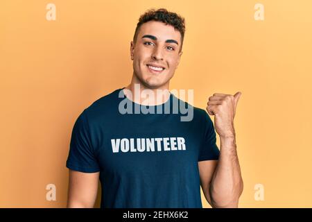 Un jeune homme hispanique portant un t-shirt de volontaire pointant vers le dos avec la main et les pouces vers le haut, souriant confiant Banque D'Images