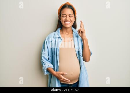 Belle femme hispanique s'attendant à un bébé, touchant le ventre enceinte gestante doigt croisé souriant avec espoir et les yeux fermés. Chance et superstitititieux Banque D'Images