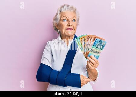 Femme sénior à cheveux gris portant le bras sur une élingue tenant des billets de banque en dollars australiens souriant regardant sur le côté et regardant loin de penser. Banque D'Images