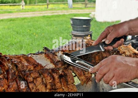 Gros plan des mains de l'homme coupant un morceau de cochon de coq (barbecue) dans le jardin. Banque D'Images