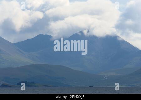 La route lointaine au-dessus de l'eau serpente vers une énorme montagne rocheuse sur une île écossaise. Des nuages lumineux baignés de soleil couvrent le sommet de la montagne. Paysage. Banque D'Images