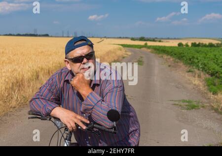 Portrait extérieur d'un homme barbu, chubby senior se pencha sur des coudes pour manipuler la barre de son vélo lors d'un court repos sur une route de campagne en été Banque D'Images