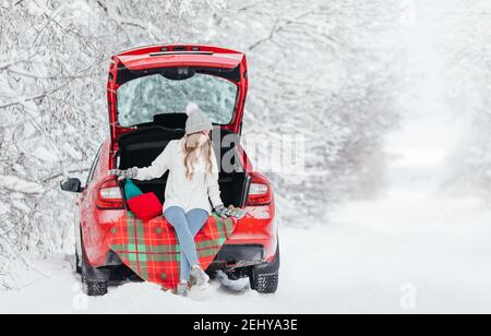 Une jeune femme dans un chandail chaud, un chapeau et des mitaines est assise sur un carreaux dans le tronc d'une voiture rouge sur le fond d'une forêt d'hiver Banque D'Images