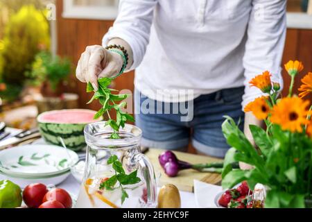 Femme à la main mettant la feuille de menthe dans la limonade froide dans la carafe. Préparation de la boisson pour la fête du jardin. Main féminine tenant les herbes sur la verseuse en verre Banque D'Images