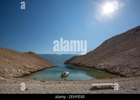 Une petite crique sur Pag avec un bateau amarré et un banc sur la plage entouré de falaises surplombant la mer bleue, le ciel et le soleil Banque D'Images