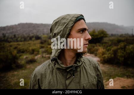 Jeune athlète masculin de forme se reposant tout en se concentrant sur la montagne de gravier chemin devant avec des buissons et des collines derrière lui Banque D'Images