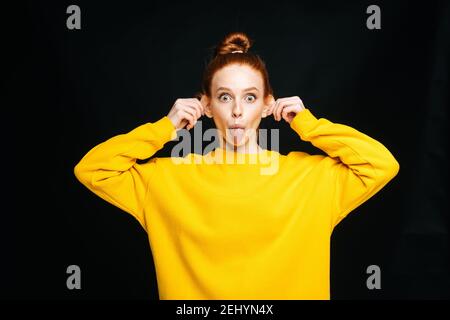 Folle joyeuse jeune femme en chandail jaune touchant les oreilles avec mains et la langue Banque D'Images