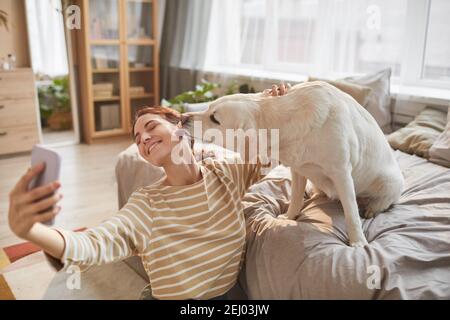 Portrait ton chaud de la jeune femme heureuse prenant le selfie avec le chien donnant des baisers dans l'intérieur confortable de la maison éclairé par la lumière du soleil, l'espace de copie Banque D'Images