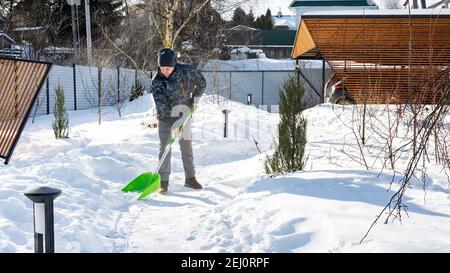 Les conséquences de fortes chutes de neige dans la cour. Un homme nettoie la neige des pistes à l'aide d'une grande pelle par beau temps d'hiver. Le jardinier prend c Banque D'Images