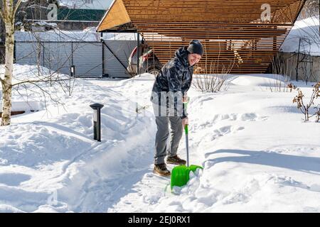 Un ouvrier nettoie la neige des chemins du jardin avec une pelle par temps ensoleillé. Entretien du jardin en hiver. D'énormes dérives de neige après un blizzard. La cour était Banque D'Images