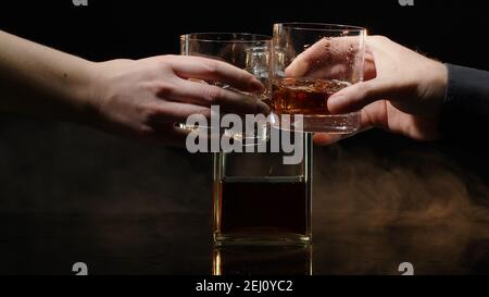 Deux mains avec des verres de whisky cognac et des glaçons pour des encouragements sur fond noir. Célébration de la réussite d'affaires, Noël, anniversaire, anniversaire. Toasts avec verres de bourbon, rhum Banque D'Images