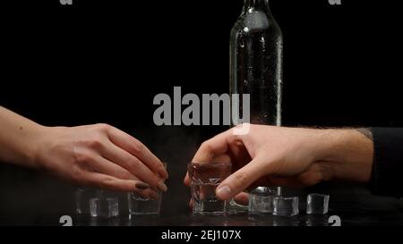 Barman déversant deux doses de vodka avec des glaçons dans une bouteille dans des verres sur fond noir. Deux mains avec des verres de vodka faisant Santé. Porter des toasts avec des verres de tequila ou du saké Banque D'Images