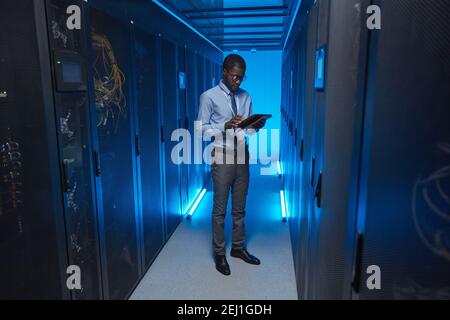 Portrait complet d'un homme afro-américain debout près d'une armoire de serveur tout en travaillant avec un superordinateur dans un centre de données et en tenant une tablette, un espace de copie Banque D'Images