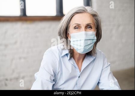 Portrait d'une femme aînée aux cheveux gris portant un masque facial, regardant la caméra, prise de vue d'une femme médecin thérapeute sur le lieu de travail Banque D'Images