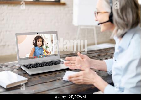 Femme mature à cheveux gris enseignant dans le micro-casque regardant l'écran, enseignant jeune femme, ayant cours virtuel avec l'étudiant, session vidéo en ligne ou Banque D'Images