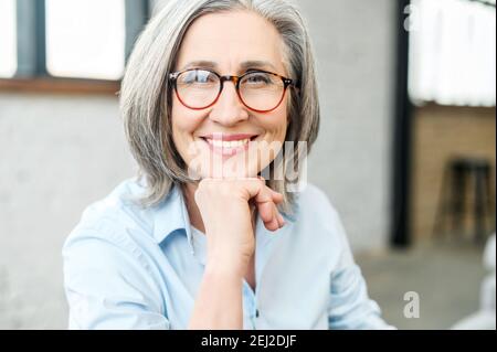 Portrait tête d'une charmante femme mûre et élégante avec sa main sur le menton, portant des lunettes et regardant l'appareil photo. Gros plan d'un milieu Banque D'Images
