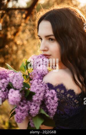 Belle jeune femme Brunette avec bouquet de lilas. Photo extérieure de la belle jeune femme avec des fleurs violettes sur fond de coucher de soleil. Fleur de printemps Banque D'Images