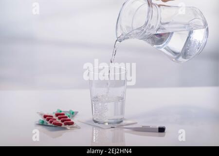 Gros plan d'une main versant de l'eau dans un verre entouré de capsules de médicaments cloques et d'un thermomètre numérique. Concept de soins de santé. Banque D'Images