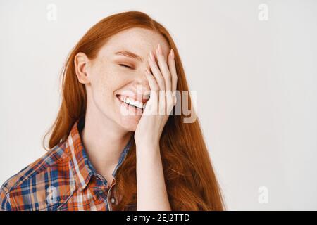 Beauté. Gros plan de la jeune femme heureuse avec des cheveux rouges naturels et une peau pâle, souriant joyeusement et couvrant la moitié du visage, debout sur fond blanc Banque D'Images