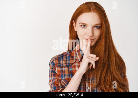 Belle femme souriante avec de longs cheveux au gingembre et des yeux bleus, partageant un secret, se précipitant avec le doigt sur les lèvres, se tenant sur fond blanc Banque D'Images