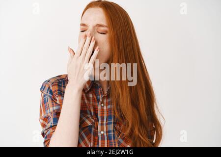 Image d'une jeune femme fatiguée avec des cheveux longs rouges et une peau pâle, couvrant la bouche tout en bâillant, se tenant épuisé sur fond blanc Banque D'Images