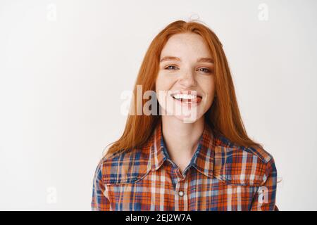 Jeune fille à tête rouge attrayante montrant la langue, souriant avec des dents blanches, regardant sans souci à l'appareil photo, debout sur fond blanc Banque D'Images