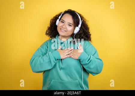 Jeune fille afro-américaine portant des vêtements de gym et utilisant des écouteurs souriant avec ses mains sur sa poitrine et geste reconnaissant sur son visage. Banque D'Images