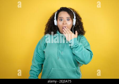 Jeune fille afro-américaine portant des vêtements de gym et utilisant des écouteurs couvrant la bouche avec la main, choqué et peur de l'erreur. Expression surprise Banque D'Images