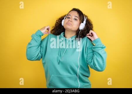 Jeune fille afro-américaine portant des vêtements de sport et utilisant des écouteurs tenant son t-shirt avec une expression réussie Banque D'Images