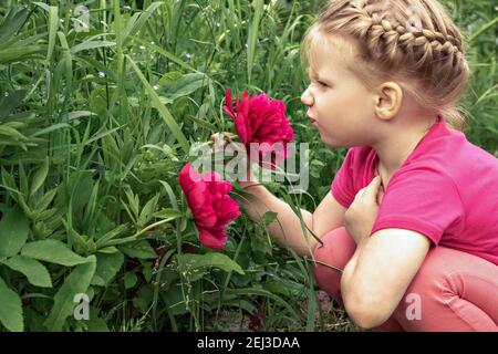 Une petite fille s'assoit près d'un lit de fleur dans le jardin et sniffs pivoines rose vif Banque D'Images