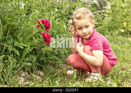 La petite fille est perdue dans la pensée, assise par le lit de fleur dans le jardin des pivoines roses. Banque D'Images