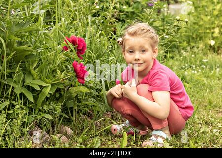 Une petite fille est assise souriante à un lit de fleurs dans le jardin de pivoines roses. Banque D'Images