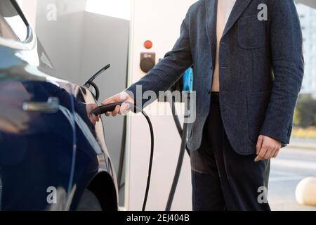 Image rognée d'un jeune homme en tenue d'affaires, en chargeant sa voiture électrique moderne à la station de charge de véhicule électrique extérieur Banque D'Images