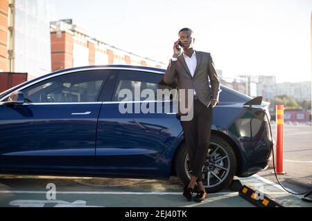 Concept de charge de voiture électrique. Portrait complet d'un jeune homme beau à la peau sombre, debout à la station de charge et parlant téléphone whie Banque D'Images