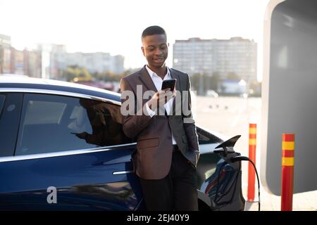 Vue de face d'un jeune homme africain heureux et agréable en treandy tenue formelle, se sentant détendu, en utilisant le téléphone tout en rechargeant sa voiture électrique de luxe moderne à Banque D'Images