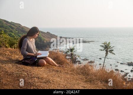 Jolie femme asiatique est assise sur le bord de la montagne avec une vue sur la mer, la lecture d'un livre. Plan d'ensemble Banque D'Images