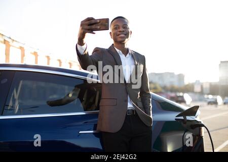 Portrait d'un jeune homme noir élégant en costume d'affaires, faisant le selfie photo sur son smartphone tout en s'appuyant sur sa voiture électrique, en chargeant la batterie Banque D'Images