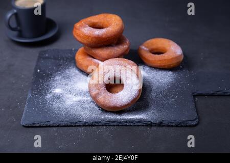 Beignets avec sucre en poudre et une tasse de café. Beignets traditionnels en forme d'anneau frits à l'huile, sur fond noir. Nourriture à la poubelle. Gros plan. Banque D'Images