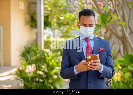 Homme d'affaires africain avec masque médical chirurgical pour la protection et l'utilisation téléphone mobile extérieur Banque D'Images