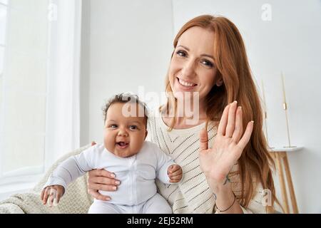 Bonne mère agitant la main tenant bébé fille drôle regardant l'appareil photo à la maison. Banque D'Images