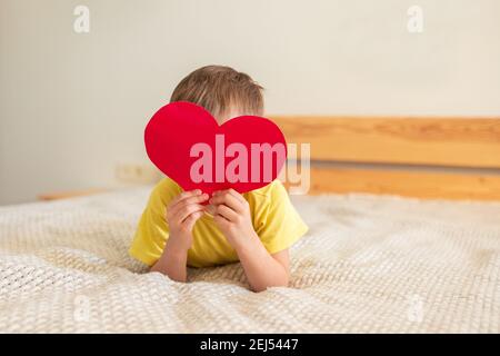 Petit garçon allongé sur le lit et tenant un coeur rouge en papier coloré, couvrant son visage. Concept pour le 8 mars, Fête des mères. Carte postale Banque D'Images