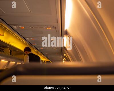 Stewart debout dans l'allée d'une cabine d'avion. Porte-bagages suspendue ouverte et voyants jaunes allumés. Vue depuis le siège passager. Banque D'Images