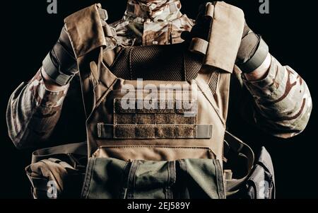 Photo d'un soldat en uniforme de camouflage et gants tactiques tenant une veste blindée militaire sur fond noir. Banque D'Images