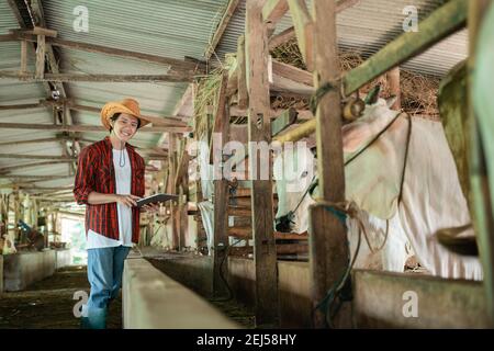 un fermier souriant portant des vêtements décontractés se tient debout tout en utilisant une tablette numérique en arrière-plan de la ferme de bétail Banque D'Images
