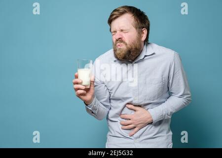 Intolérance au lactose. Un homme tient un verre de lait dans sa main et souffre de douleurs abdominales sévères. Arrière-plan bleu. Banque D'Images